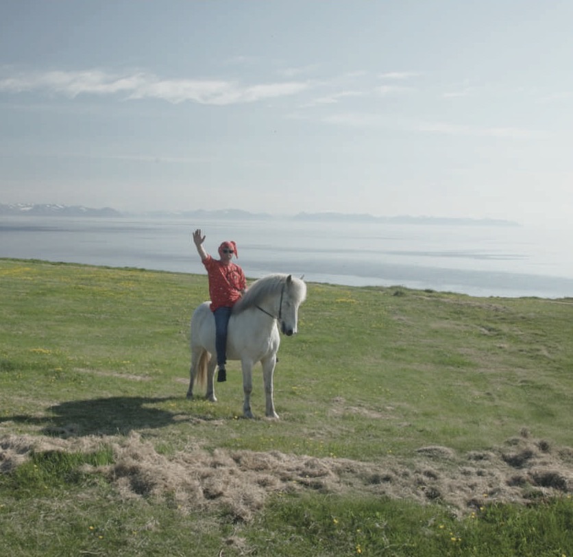 A rider waves from a white horse on a clifftop above the sea